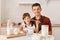 Smiling male with dark hair wearing maroon shirt and black apron sitting in kitchen with his cute female child with corolla in