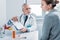 smiling male doctor with stethoscope over neck pointing by pencil at clipboard to female patient at table