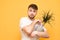 Smiling man with a beard stands on a yellow background, holding a flowerpot with a plant in his hands. Adult man wears a white T-