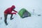 Smiling man dressed red warm mountaineering down jacket making the protective wall from snow bricks around camp tent on glacier