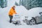 Smiling man in funny nepalese hat removing snow from car hood using big snow brush. The automobile freezes out on the countryside