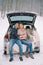 Smiling man and woman sit hugging in car trunk on blankets under snowfall in forest