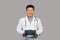 Smiling mature chinese man therapist in white coat with stethoscope use tablet, isolated on gray background