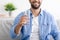 Smiling middle aged caucasian guy with beard sits on sofa and holds glass with clean still water