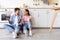 Smiling millennial european woman and man with cups of drink, sitting on floor, talk in kitchen interior