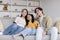 Smiling millennial korean mother, father and teen girl hugging and watch tv, sit on sofa in living room