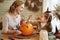 Smiling mother and child girl drawing scary faces on Halloween pumpkins while sitting in cozy kitchen