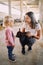 Smiling mother, squatting down, shows her little daughter a black goatling in her arms