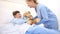 Smiling nurse takes care of a child lying in hospital bed making him a surprise by giving him a teddy bear