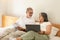 Smiling old caucasian husband and wife lie on bed, pay for bills, taxes in bedroom interior