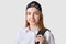 Smiling positive school girl looks directly at camera, poses isolated over white background, holding black bagpack with one hand,