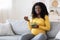 Smiling pregnant black woman eating fresh salad, sitting on sofa