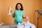 Smiling pregnant woman holds steam iron and shows ironed clean baby clothes, standing at ironing board.