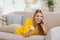 Smiling pretty millennial blond caucasian woman in yellow clothes speaks by phone, lies on sofa