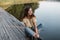 Smiling pretty woman with curly hairs in a yellow fashionable blouse with vintage jeans is resting on a wooden pier by the lake in