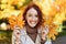 Smiling pretty young european red-haired lady in raincoat hold yellow leaves, has fun alone in city park