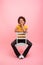 Smiling pretty young mixed race woman with curly hair in casual sits on chair,  on pink background