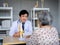 Smiling professional Asian man orthopedic doctor in white suit pointing to knee joint anatomy model to explain for senior female.