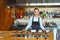 Smiling proud waitress woman standing behind counter looking at camera - Portrait of happy beautiful confident female with apron