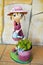 Smiling puppet made in colorful steel that shows a happy cross-eyed girl watering can a bucket with a green plant on a yellow