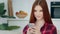 Smiling red-haired caucasian girl model woman sits in kitchen room holds modern gadget smartphone looks at mobile