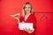 Smiling satisfied and proud blonde woman look at camera in red shirt with box of shoes in hands on red background.