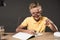 smiling schoolboy in eyeglass pointing by finger and doing homework at table with books colour pencils and textbook on grey