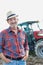 Smiling senior farmer wearing hat, hands in pocket while standing against mature farmer driving tractor in field