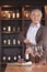 Smiling senior man opening wine bottle and looking at camera, shelf with wine in the background