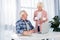 smiling senior man sitting at table with laptop and looking at wife with cups of coffee