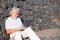 Smiling senior white-haired man sitting outdoor on staircase using laptop computer. Relaxed elderly man wearing necklace