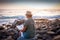 Smiling senior woman with hat sitting on the pebble beach reading a book. Positive moment of relax for the retired grandparent.