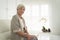 Smiling senior woman sitting on sofa and looking at camera. Awaken old woman with grey hair and pajamas in the early morning light