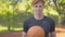 Smiling sportsman posing in sunlight on outdoor basketball court. Portrait of young confident redhead man holding ball