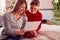 Smiling Teenage Daughter Wearing Pyjamas Looking At School Exam Report With Mother In Bedroom