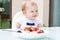 A smiling toddler baby girl sits at a table in front of a plate of porridge with berries and looks away