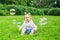 A smiling toddler sits on the grass in a summer park and looks at soap bubbles
