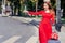 Smiling tourist girl in red dress with valise catches a taxi on street of city.