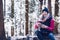 A smiling tourist is resting on a tree trunk, drinking hot tea and eating a hamburger on a mountain trail on a cold day.