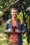 Smiling traveler with backpack and binoculars listening music in headphones in green tropical forest.