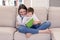 Smiling tween girl reading book to her little brother sitting in her arms with soft furry rabbit toy on sofa at home.