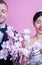 Smiling wedding couple with artificial flowers standing with eyes closed against pink background