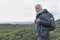 Smiling white-haired elderly man looking at camera while hiking among green bushes and the sea. Horizon over the water. Active