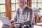 Smiling white haired senior man in winter sweater sitting in living room using laptop computer. Carefree elderly grandfather