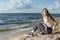 Smiling Winsome Blond Caucasian Girl in Casual Clothing Sitting While Resting In Front of Sea Shore At Daytime