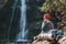 Smiling Woman with backpack dressed in active trekking clothes sitting near the mountain river waterfall and enjoying the