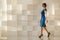 Smiling woman in blue dress walking against modern wall with wallet in hand