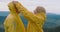 Smiling woman putting on hood of yellow rain coat on man standing on mountain peak. Side view portrait of loving mature