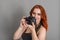 Smiling woman with red hair holds a modern camera in her hands. Gray background, creativity, photographer, beautiful