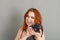 Smiling woman with red hair holds a modern camera in her hands. Gray background, creativity, photographer, beautiful